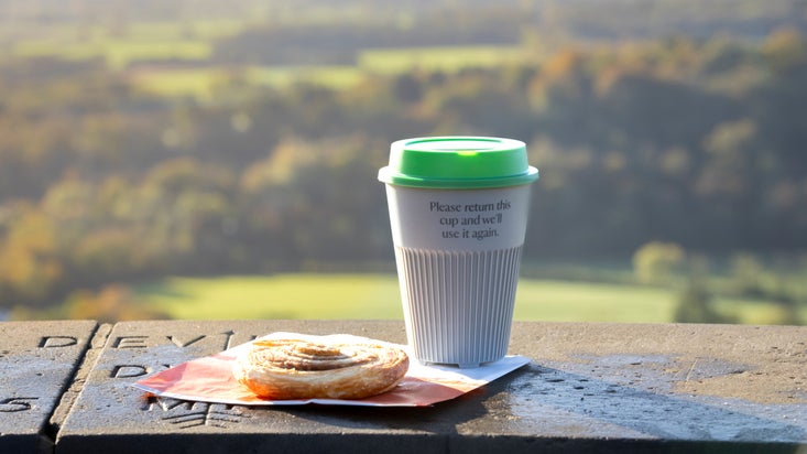 Returnable cup and a pastry on a napkin, sitting on the stone viewpoint at Box Hill with a view to the South Downs behind